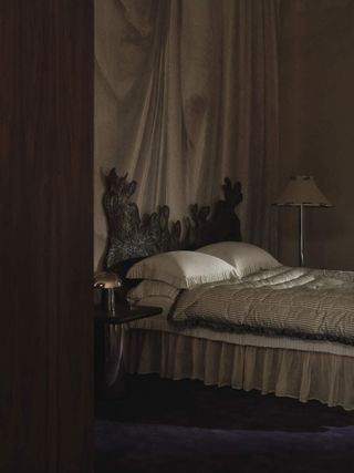 Image of a dark, moody bedroom. There is silk, light gray, and white bedding with a ruffled bed skirt, and a dark, abstract headboard. Behind the bed is off-white drapery.