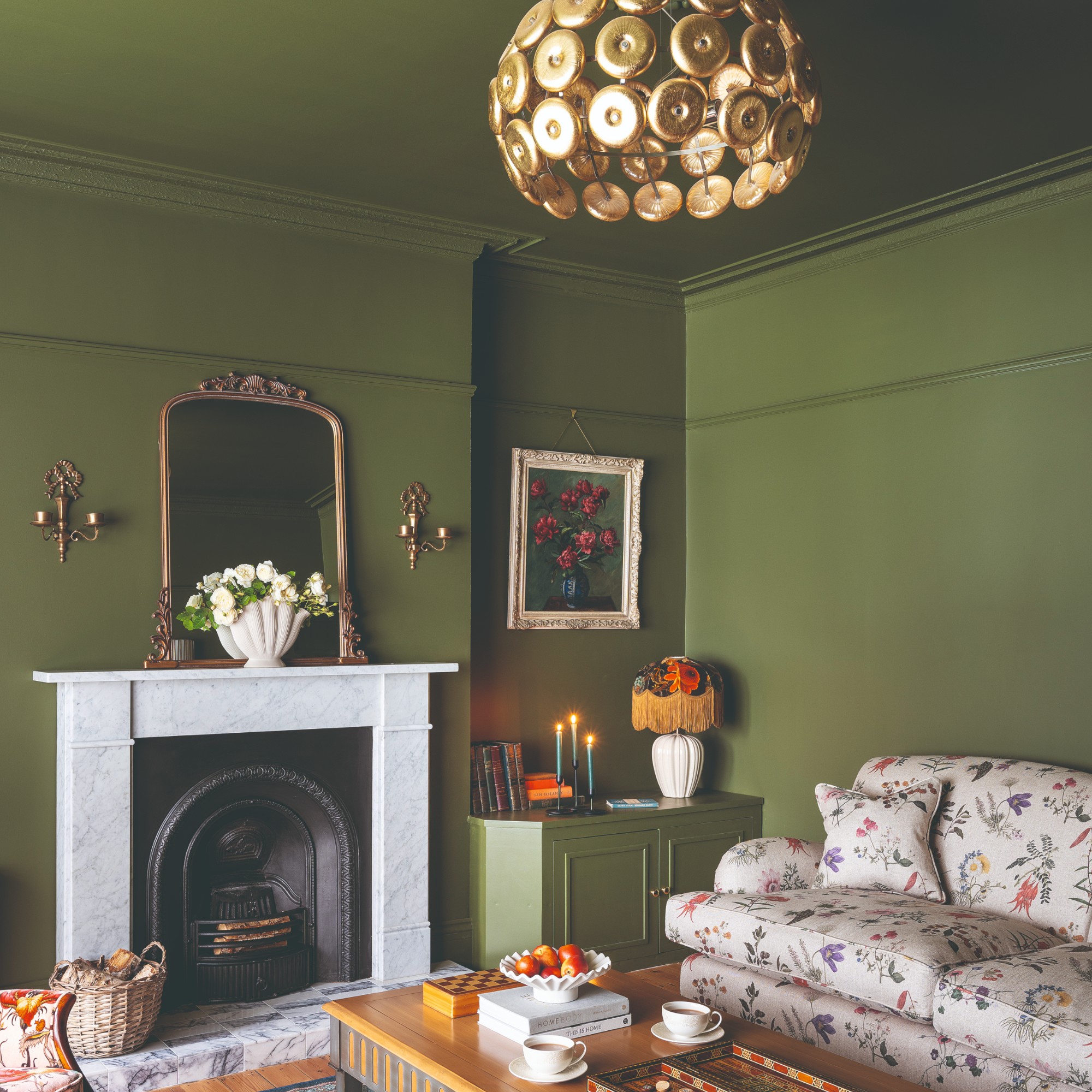 A colour-drenched green living room with a floral sofafringe-trimmed table lamp and gold ceiling light fixture