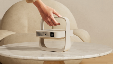 Lifestyle shot of the Xgimi Z6X 5th Generation projector unfolded on a light marble table. A woman's hand is coming from the top of the image to lift the projector up