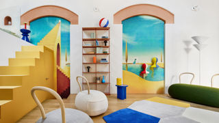 A white small apartment with a 'Mysterious Baths' mural by Owen Gent, framed by arches. In the living room are Yum Yum seats by Opinion Ciatti, a color-blocked rug, and a blue stool beside a shelf