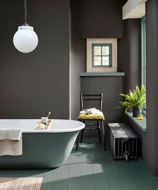 Traditional interior with bathroom painted in modern paint colors. Dark gray painted walls, soft blue used as a painted accent color around windows, flooring and on bath. Cozy alcove with small window, black wooden chair and low radiator, rounded glass pendant hanging over bath.