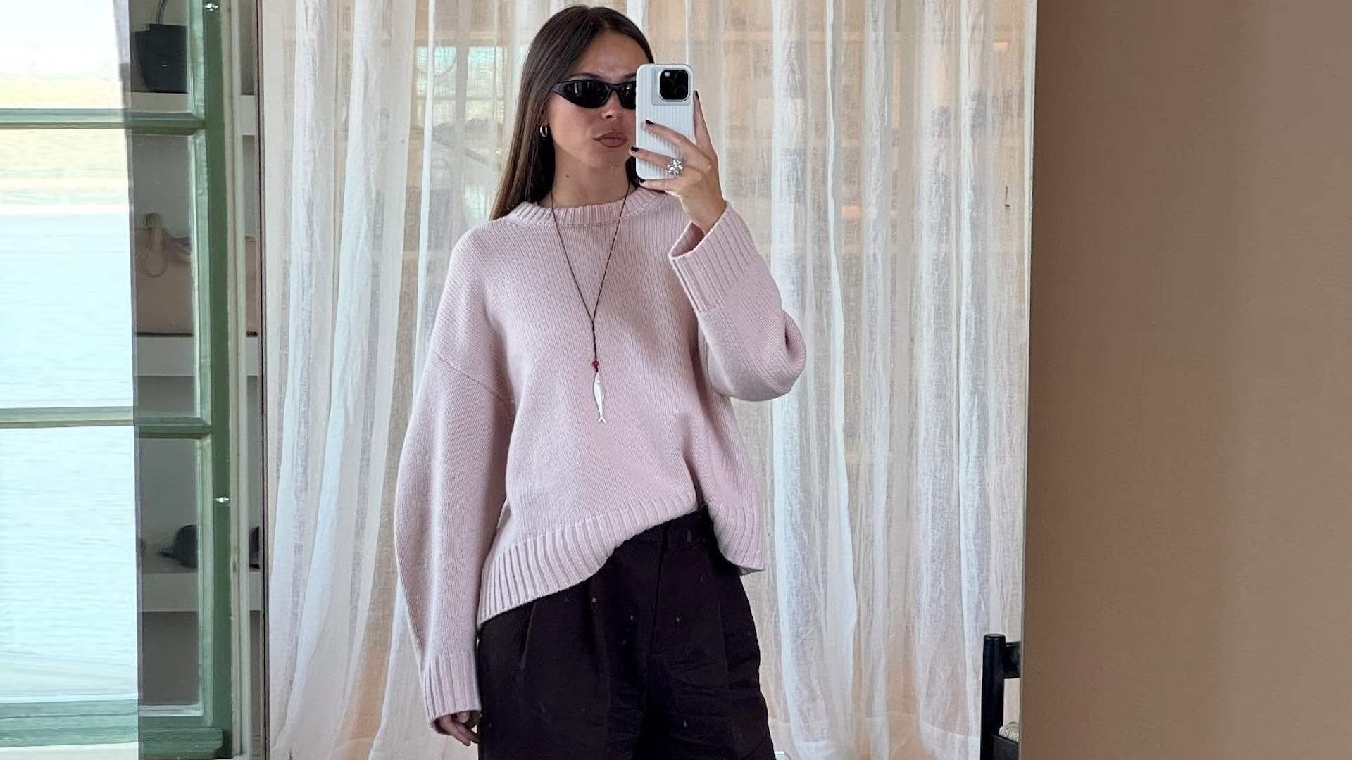 Scandinavian fashion creative Johanna Lager poses for a mirror selfie wearing black oval sunglasses, a chunky pink crewneck sweater, a red cord necklace with a silver fish pendant, and black pants