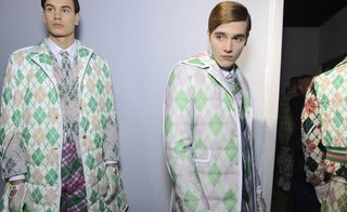 Two male models wearing looks from Moncler Gamme Bleu's collection. They are wearing shirts, ties, coats, trousers and gloves in white, green and pink with a repeating diamond pattern