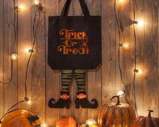 Black and orange tote bag made using Cricut machinery with witches legs. In background, orange pumpkins, wooden backdrop and LED fairy lights