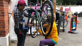 Two people working together on a bike in a stand outdoors. The person on the left holds the front wheel at an angle while the person on the right kneels and checks for play in the hubs.