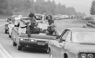 A couple play the guitar sitting on their car on the way to the Woodstock Festival,