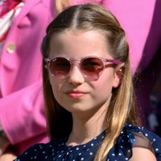 Princess Charlotte wearing a blue polka dot dress and pink sunglasses smiling with her lips closed on a sunny day