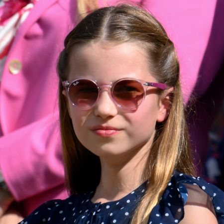 Princess Charlotte wearing a blue polka dot dress and pink sunglasses smiling with her lips closed on a sunny day