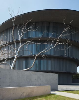 A closer look at the exterior of the He Art Museum. The building is circular with concrete walls in front of it and a tree with naked branches.
