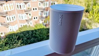 The Sonos Era 100 wireless speaker, pictured on a balcony above green foliage.