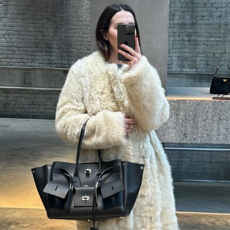 Caitlin Burke wearing a white shearling coat and carrying a black Balenciaga Bel-Air Carryall bag with silver hardware. 