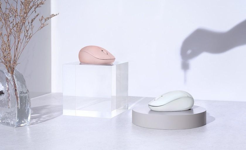 Two contoured wireless computer mice, one in dusty pink and one in mint green, are displayed on a clear acrylic block and a round grey pedestal in a brightly lit, stylized setting.