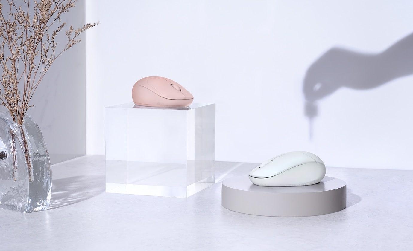 Two contoured wireless computer mice, one in dusty pink and one in mint green, are displayed on a clear acrylic block and a round grey pedestal in a brightly lit, stylized setting.