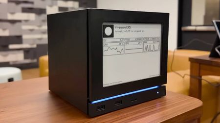 Steam Machine sitting on woodgrain table in Valve office with e-ink screen on front displaying PC performance stats.