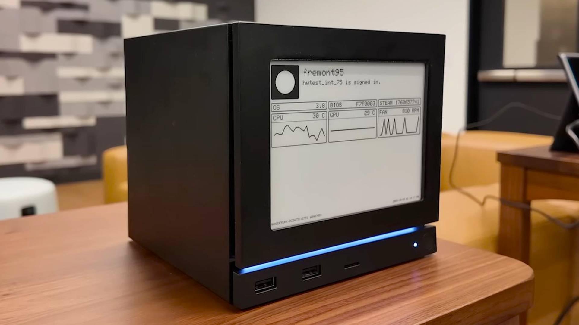 Steam Machine sitting on woodgrain table in Valve office with e-ink screen on front displaying PC performance stats.