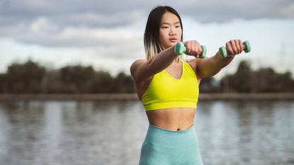 A woman performing a front raise with dumbbells as part of an outdoor workout by a lake