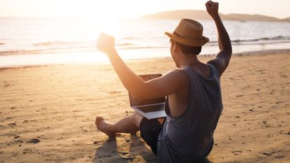 Young man holding a laptop and raising his hands in victory on a beach