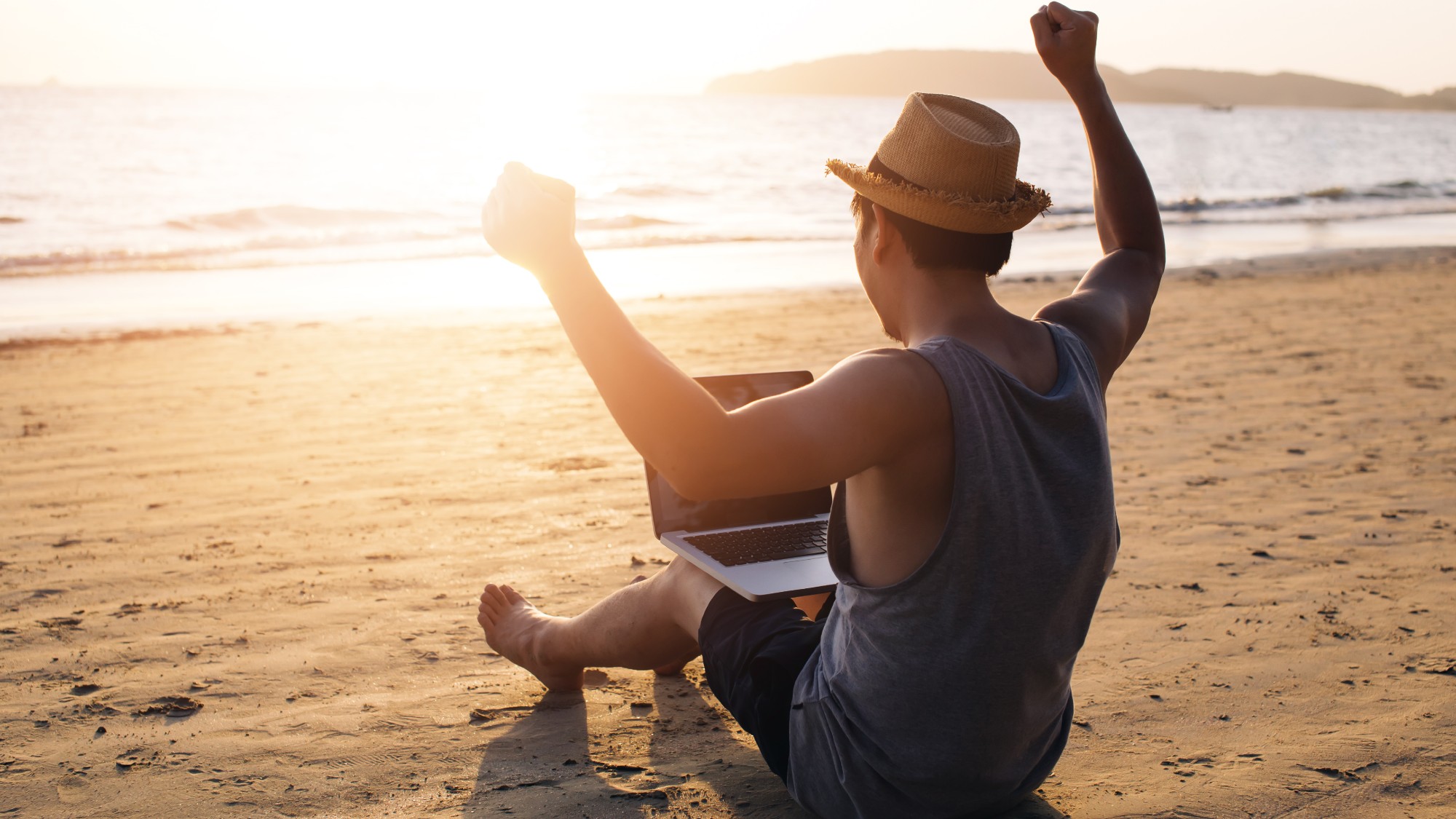 Young man holding a laptop and raising his hands in victory on a beach 