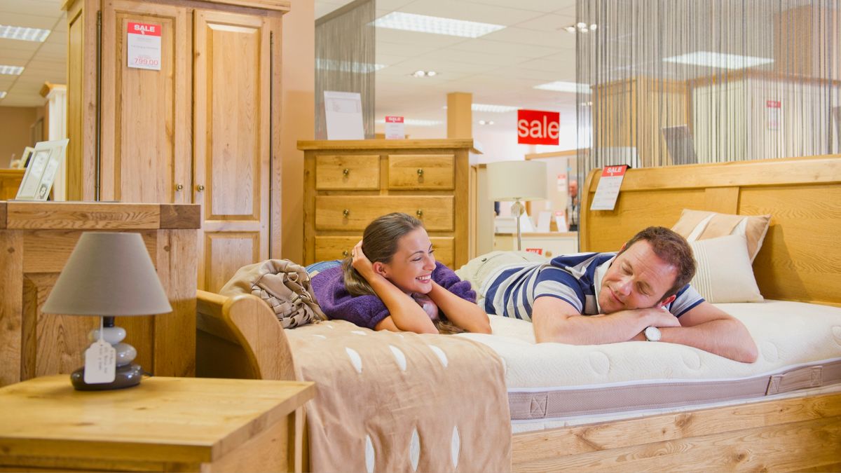 A man and woman smiling and lying on their fronts on a mattress with a for sale sign behind them