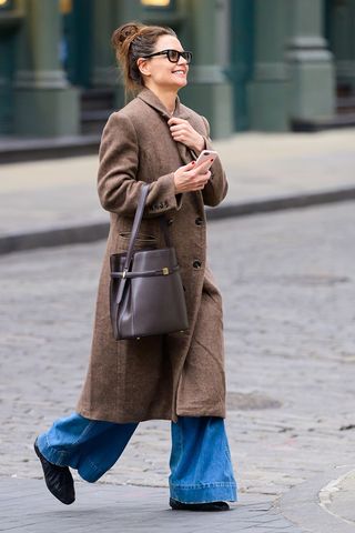 Katie Holmes wearing a brown coat, wide-leg jeans, and black ruched ballet flats in NYC.