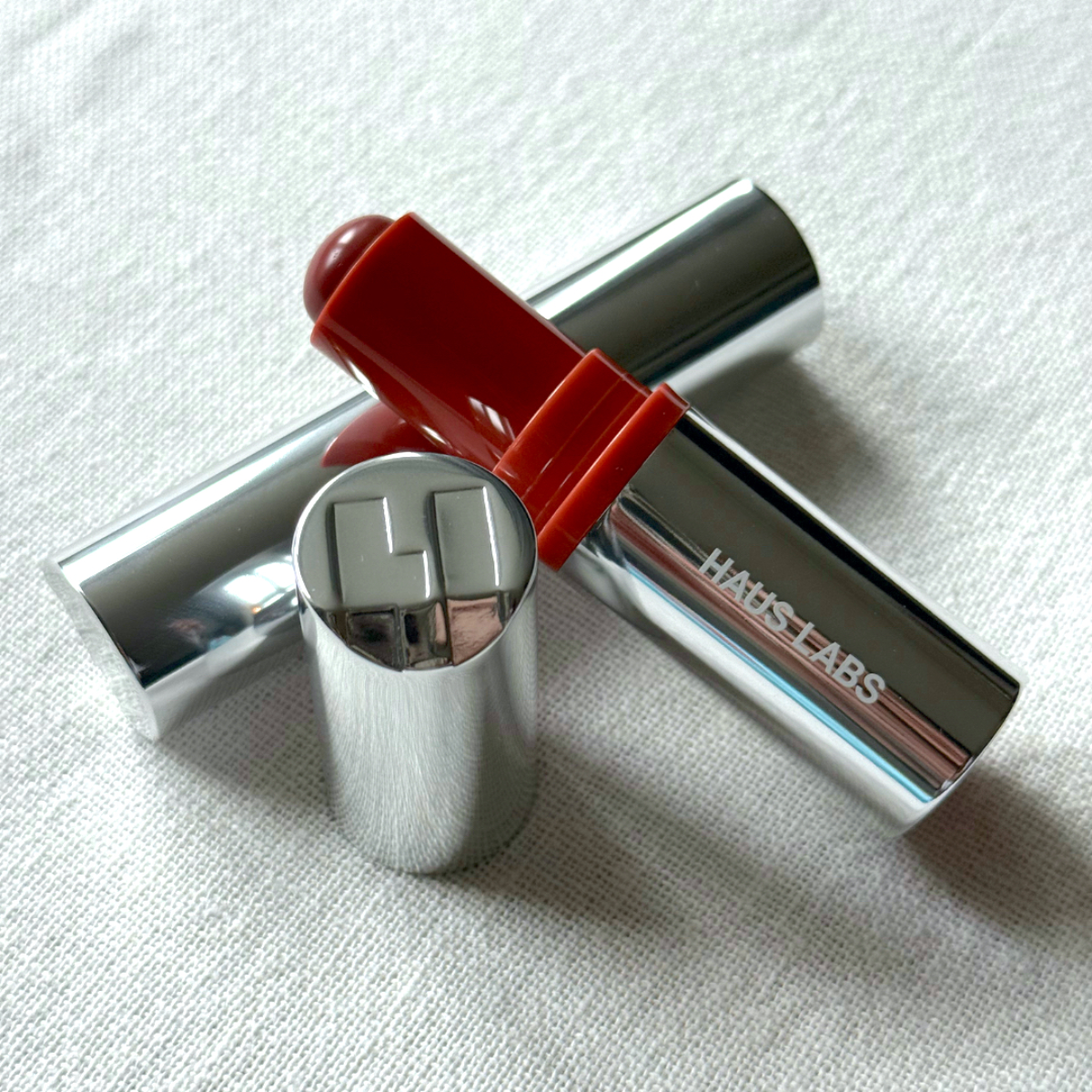 Bird&#039;s eye view of two Haus Labs Glassy Blush Balm Sticks, one with its id off, arranged in a still life ensemble on an off-white linen background