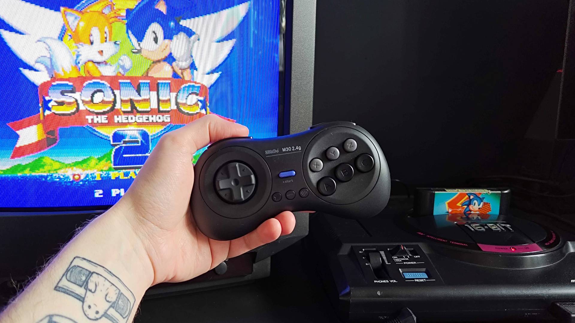 Hand holding 8Bitdo M30 Sega Mega Drive/Genesis controller with Sonic the Hedgehog 2 title on Sony Trinitron CRT TV next to Japanese console.