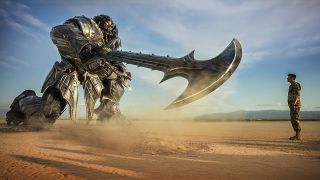 Still from the movie Transformers: The Last Knight (2017). Here we see a giant Transformer with a knight-like appearance and amor. He is down on one knee and holding forward a giant sword, pointing it towards the male soldier in front of him. In the background you can see that they are both in a flat, dry desert.