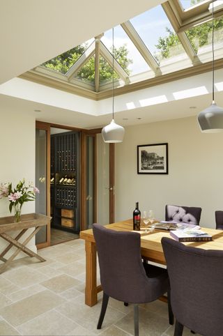 a modern country orangery interior by Westbury Garden Rooms, with a wooden table, material dining chairs, a wine room beyond and a wooden side table with accessories on top