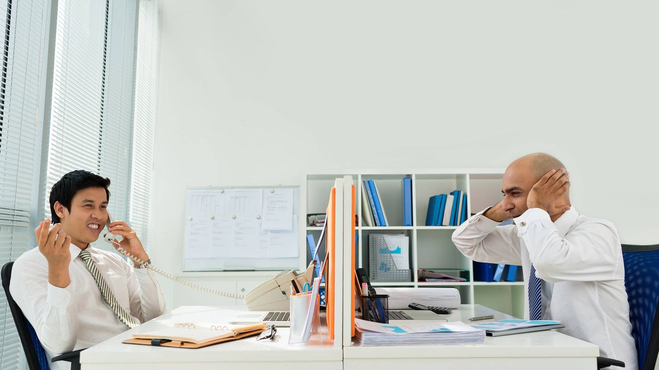 A man hold his hands over his ears as his co-worker chats on the phone nearby.