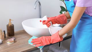 A woman cleaning a bathroom while wearing gloves, using a sponge and spray bottle