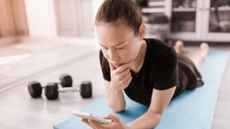 Woman lying on her front on exercise mat looking at a smartphone with two dumbbells on the floor next to her