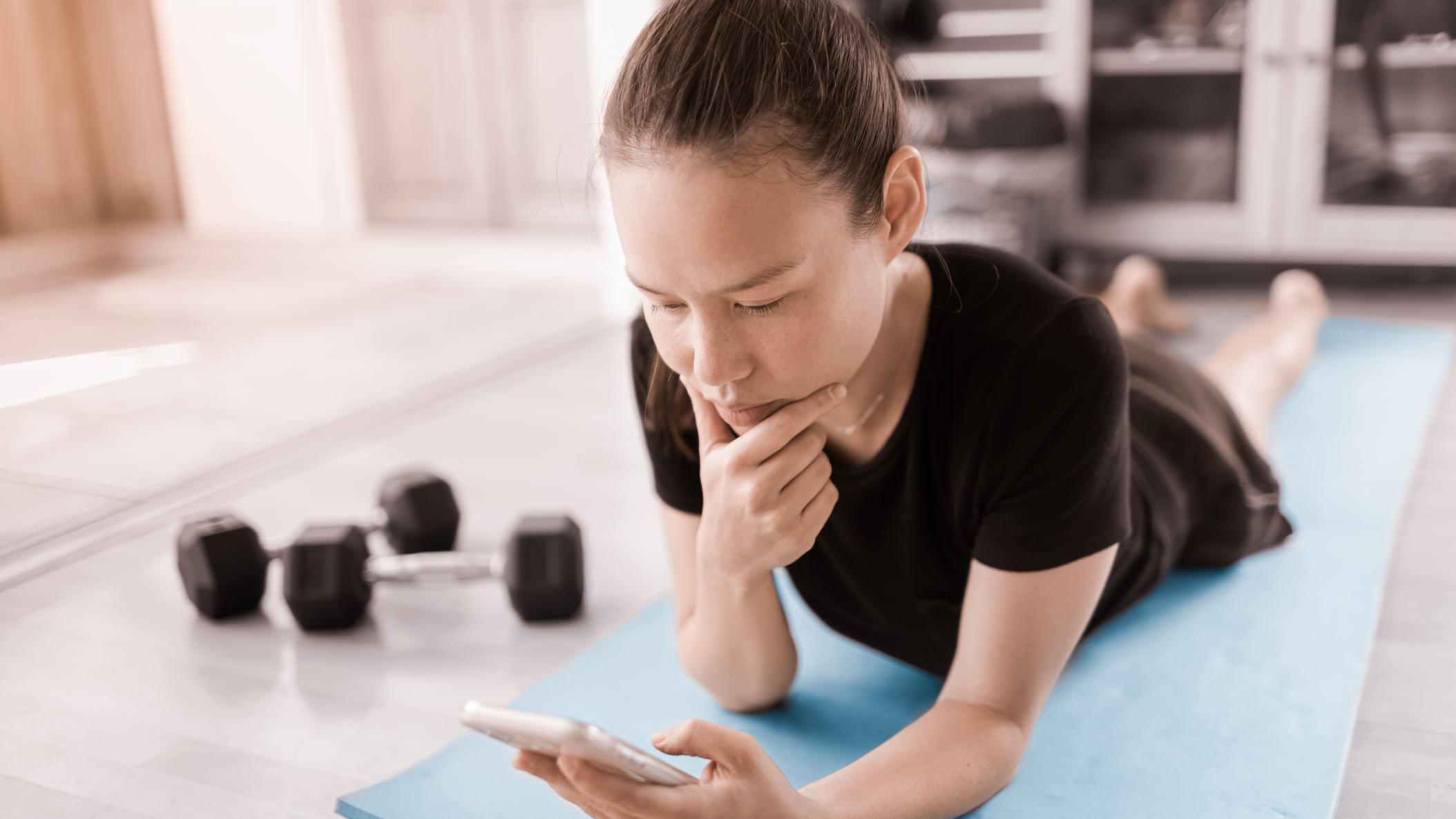 Woman lying on her front on exercise mat looking at a smartphone with two dumbbells on the floor next to her