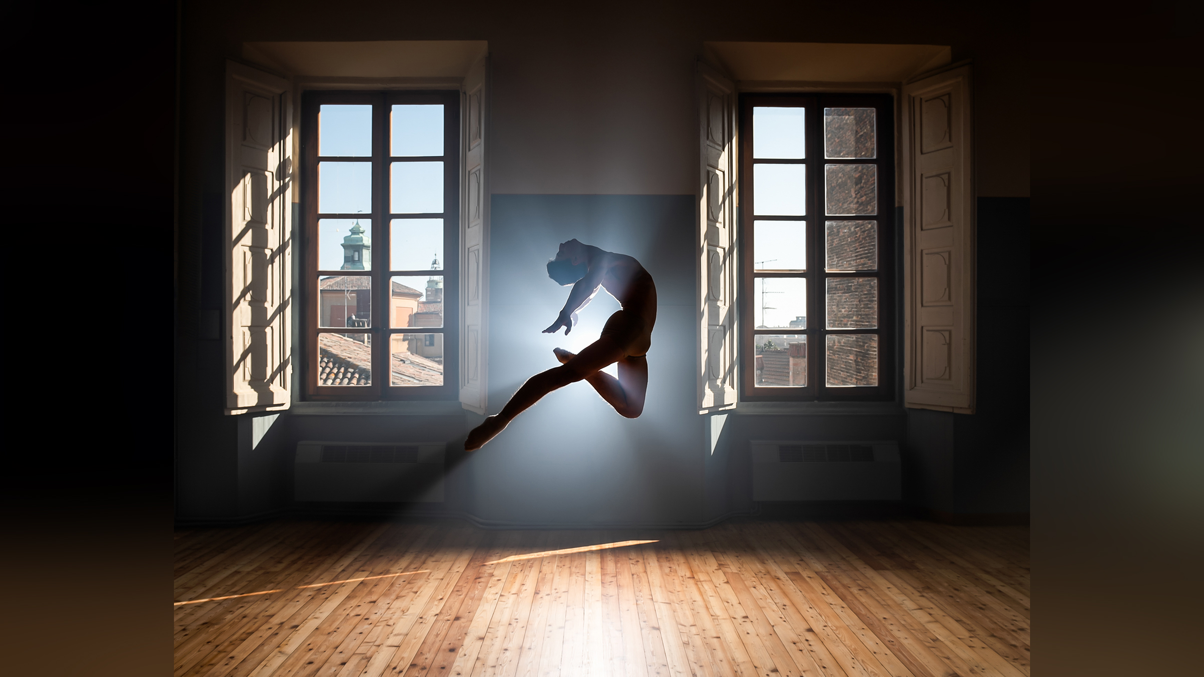 A dancer elegantly leaps in a sunlit room, framed by open windows, casting a dramatic silhouette against the bright backdrop