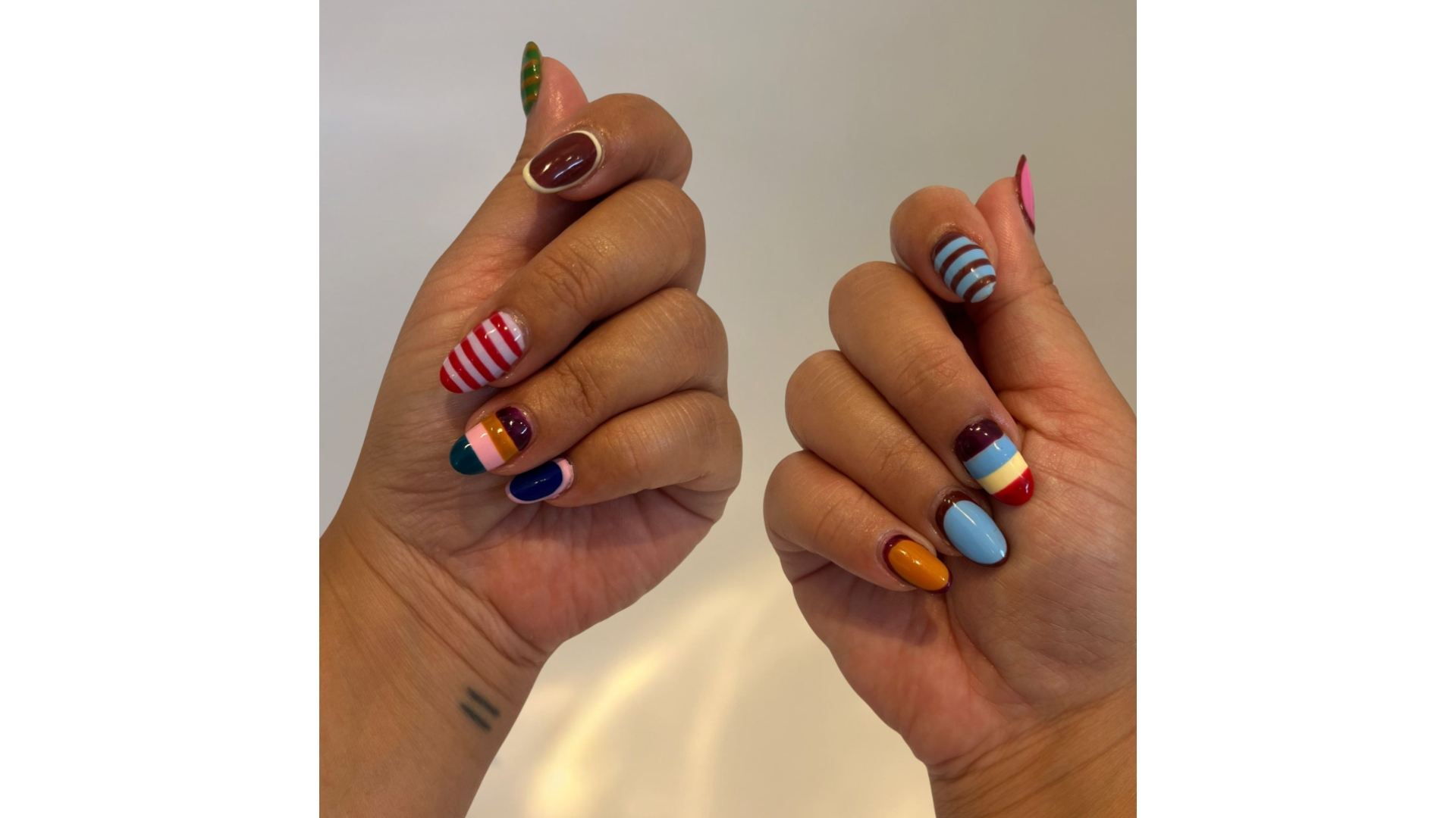 A close-up of Social Media Editor, Luisa Rossi's hands, which feature a bright, multi-coloured and maximalist, striped manicure, pictured in front of a cream background