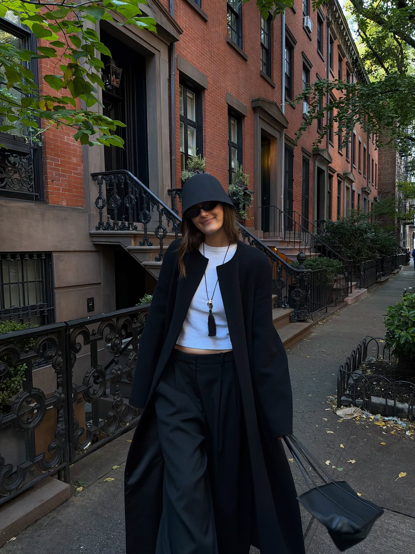 An image of @cocoschiffer wearing a collarless black trench coat, a black knit hat, a white t-shirt, a pendant necklace and a tote bag while out in New York.