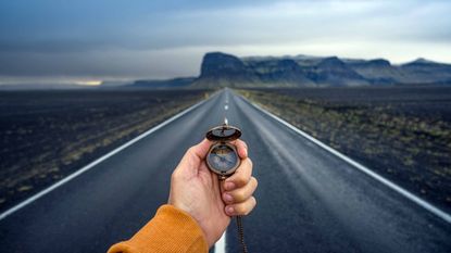 person holding compass standing on empty highway looking toward mountains