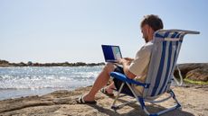 A young man working on a laptop, while sat on a beach chair at the beach, waves and clear blue sky visible in front of him.