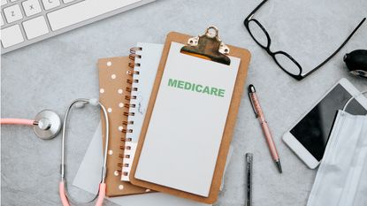 A clipboard holds a piece of paper that says Medicare on it sits among a laptop keyboard, eyeglasses and a stethoscope.