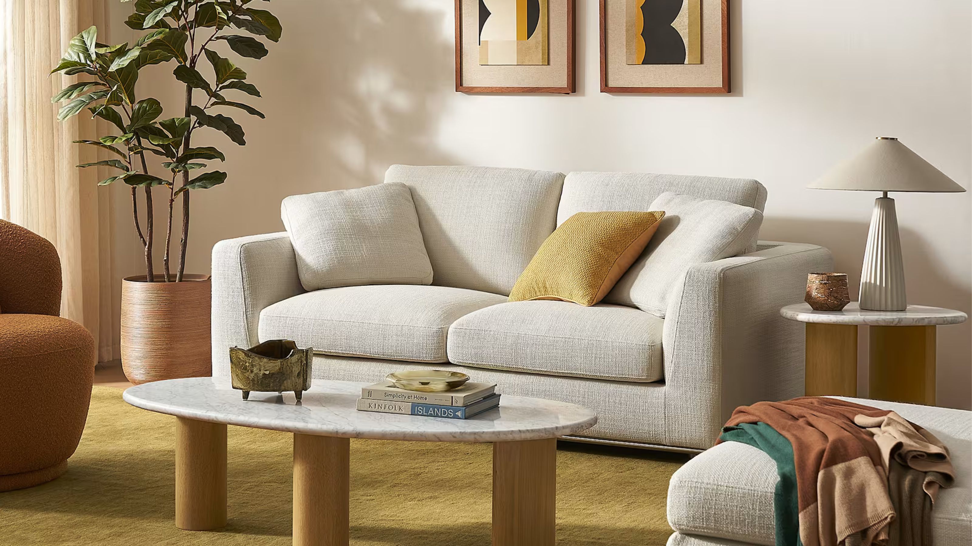 living room with white fabric sofa with yellow cushion, yellow rug, side table with white lamp, potted fiddle leaf fit, round marble coffee table with timber legs, fabric ottoman with a throw draped over it and art on the wall