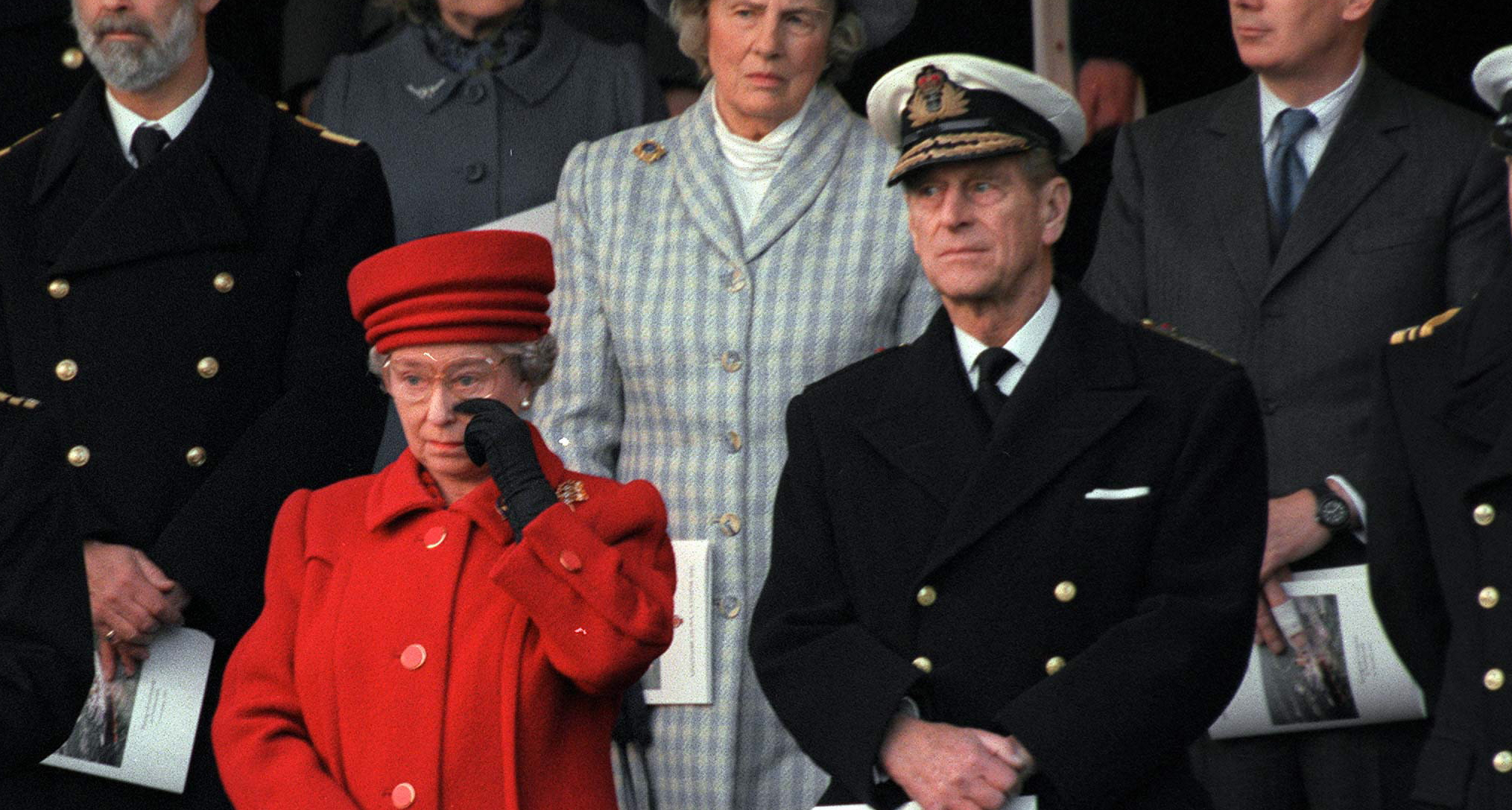 Queen Elizabeth wearing a red coat and hat wiping her eye