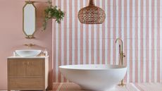 A bathroom with white and pink striped wall tiles, a white freestanding bath, and a wooden bathroom vanity