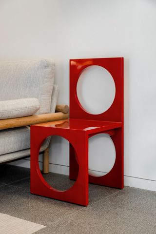 View of the 'Enzo' chair designed by hotelier Paddy McKillen - a red lacquer chair with a round cut-out design. Beside the chair is a light coloured seat with a wooden frame in a space with white walls and grey flooring