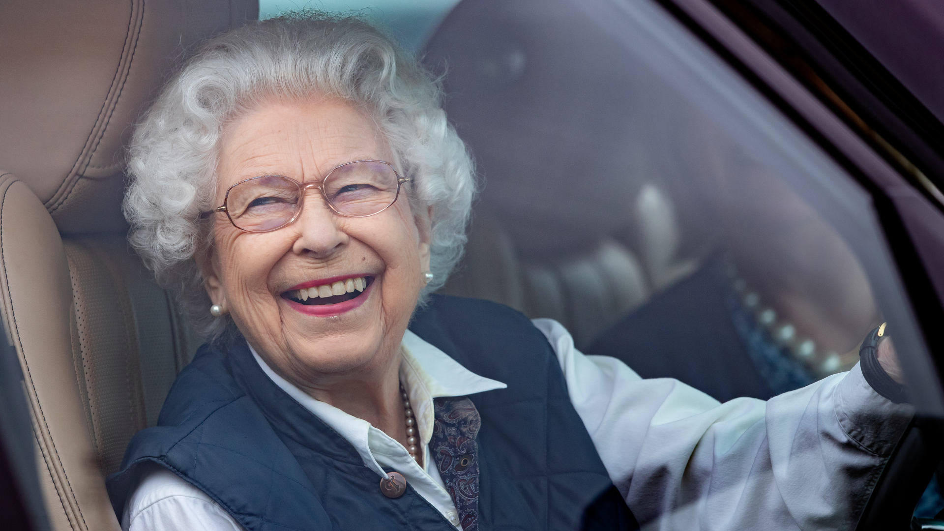 Queen Elizabeth wearing a blue vest and white shirt laughing while driving her car