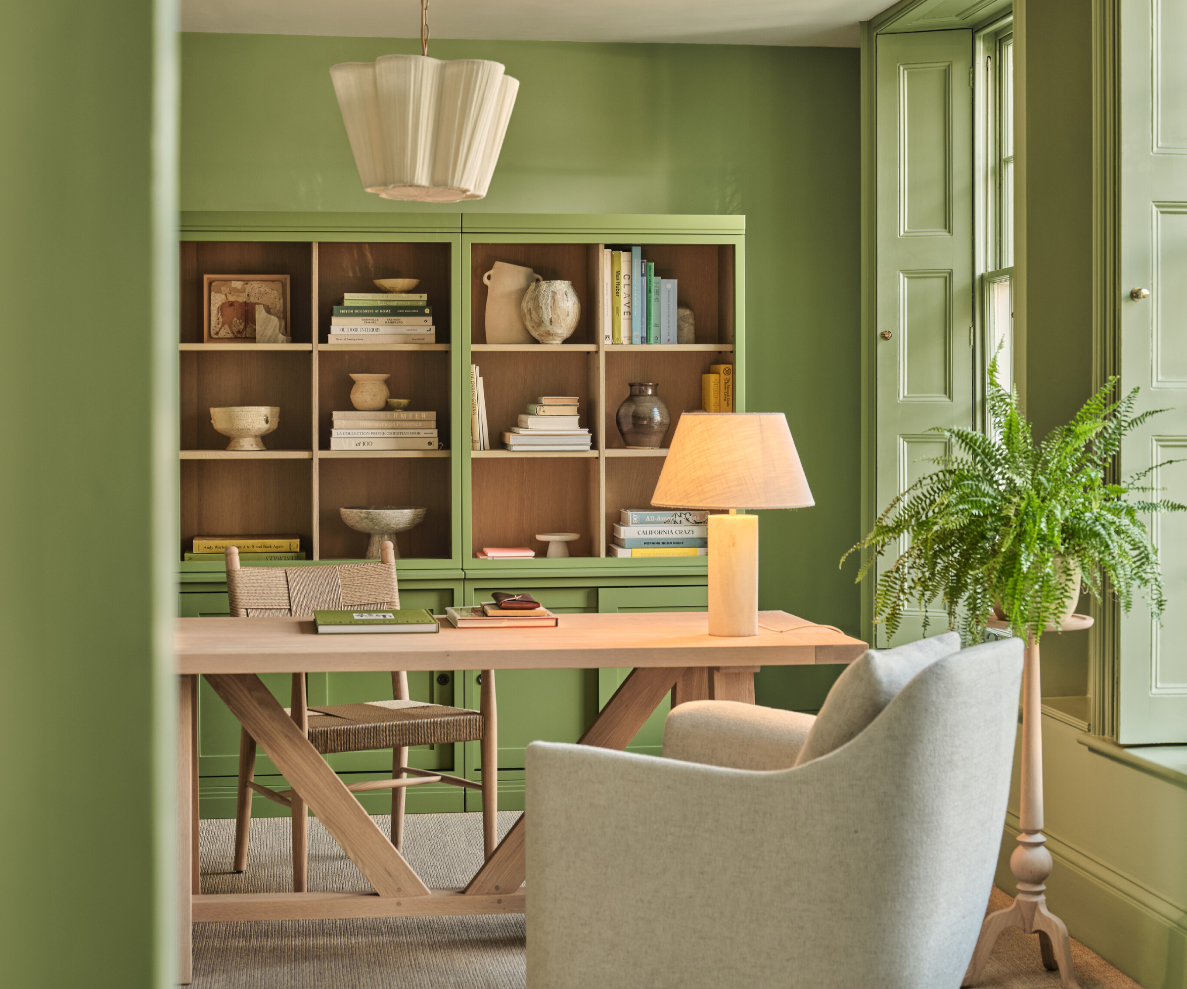 home office with green walls, window shutters, green storage with wood inside cubby holes, large wooden desk, lighting and fabric armchair