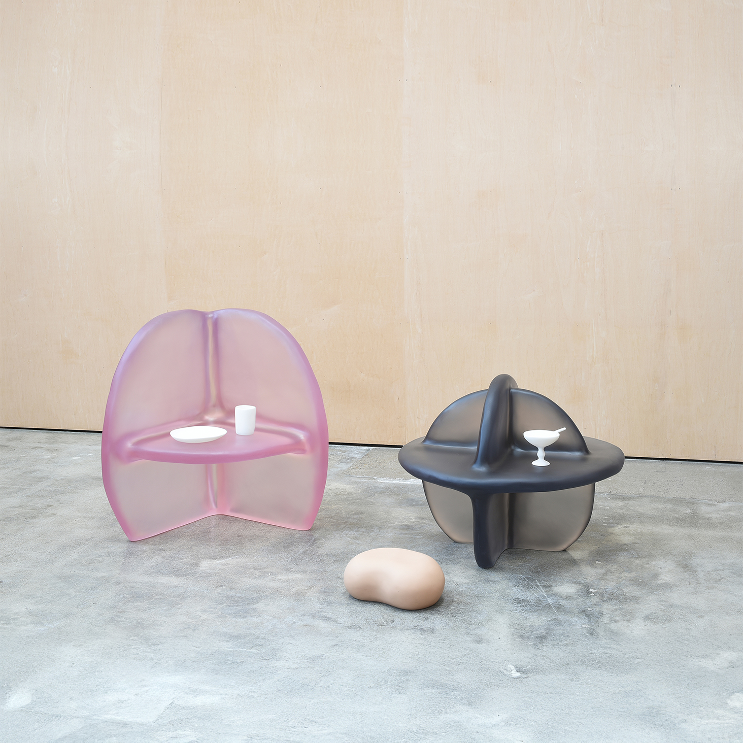 Two whimsical chairs-cum-shelves feature a blown-up, semitransparent aesthetic, one in bright pink, the other one in black, as they sit in an industrial room with cement flooring.