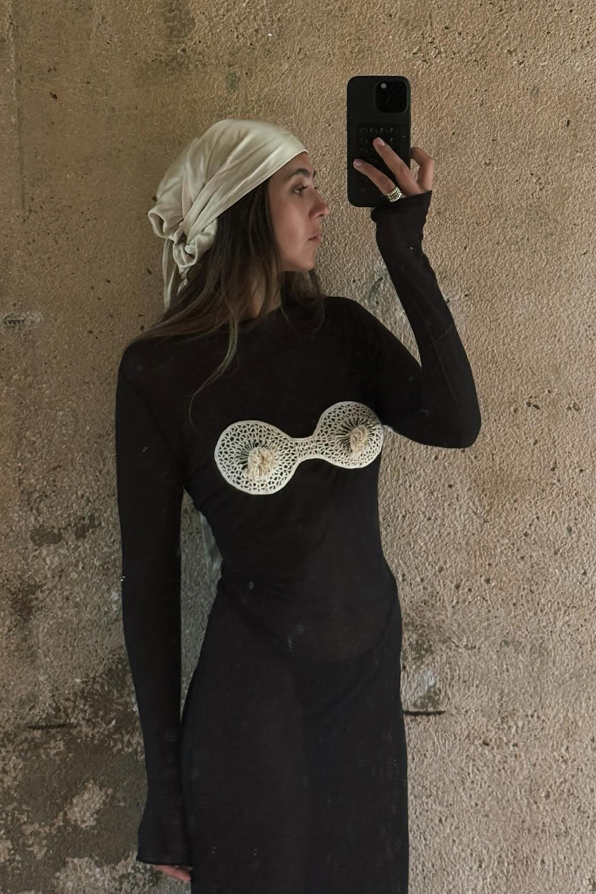 How to wear a bandana is shown in a photo of a woman standing against a wall, taking a selfie wearing a beige headscarf with a black sheer swimsuit coverup and black swimsuit