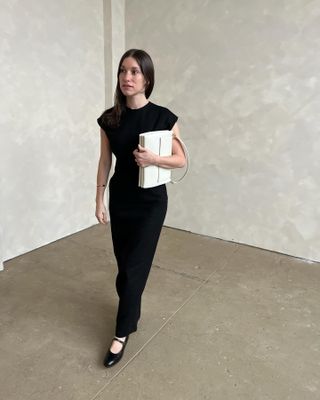 Woman wearing black dress with cap sleeves and black Mary Jane shoes, holding white structured shoulder bag, leaning against marble wall