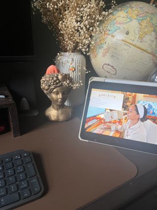 An ipad with a youtube video on, on a brown office desk, in front of an old globe, dried stems, and a gold bust with a clenaing cloth inside