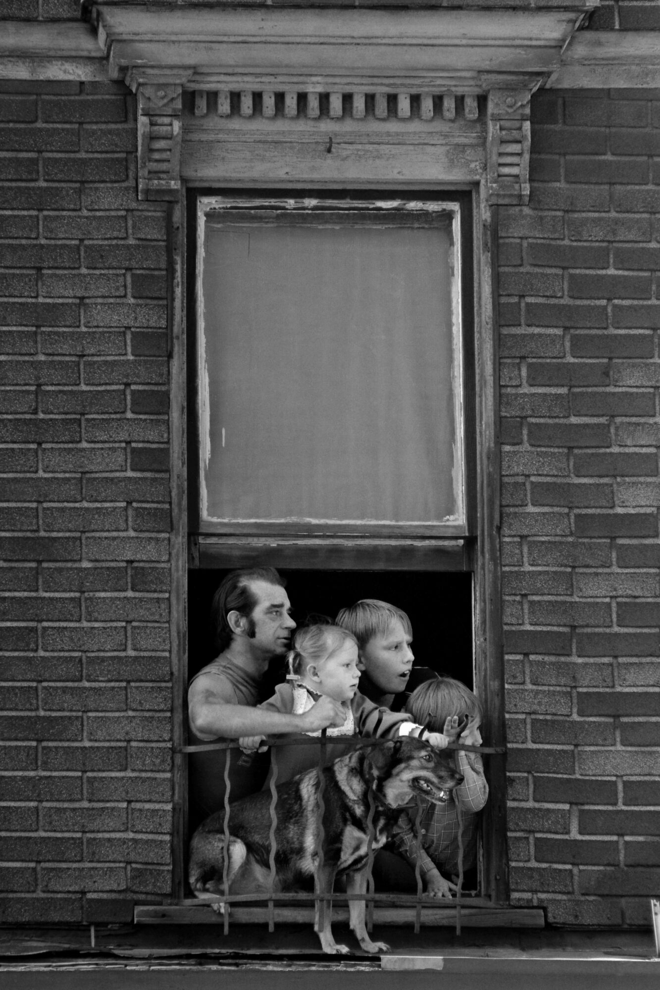 A man, three young children and a dog crowd into the open lower half of a sash window in a brick building, all looking intently at something to the right.