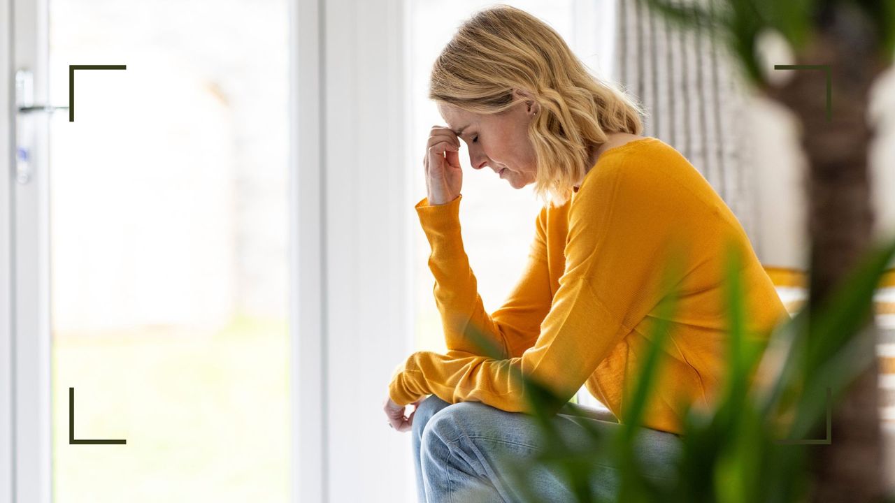 Woman holding head in hands, sitting on step next to plant at home, in pain with sinus infection